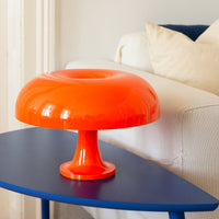 Die orange Pilzlampe steht auf einem blauen Design-Beistelltisch neben einem weißen Sofa mit cremefarbenen Kissen. Das kräftige Orange der Lampe setzt einen markanten Kontrast zur minimalistischen Einrichtung.