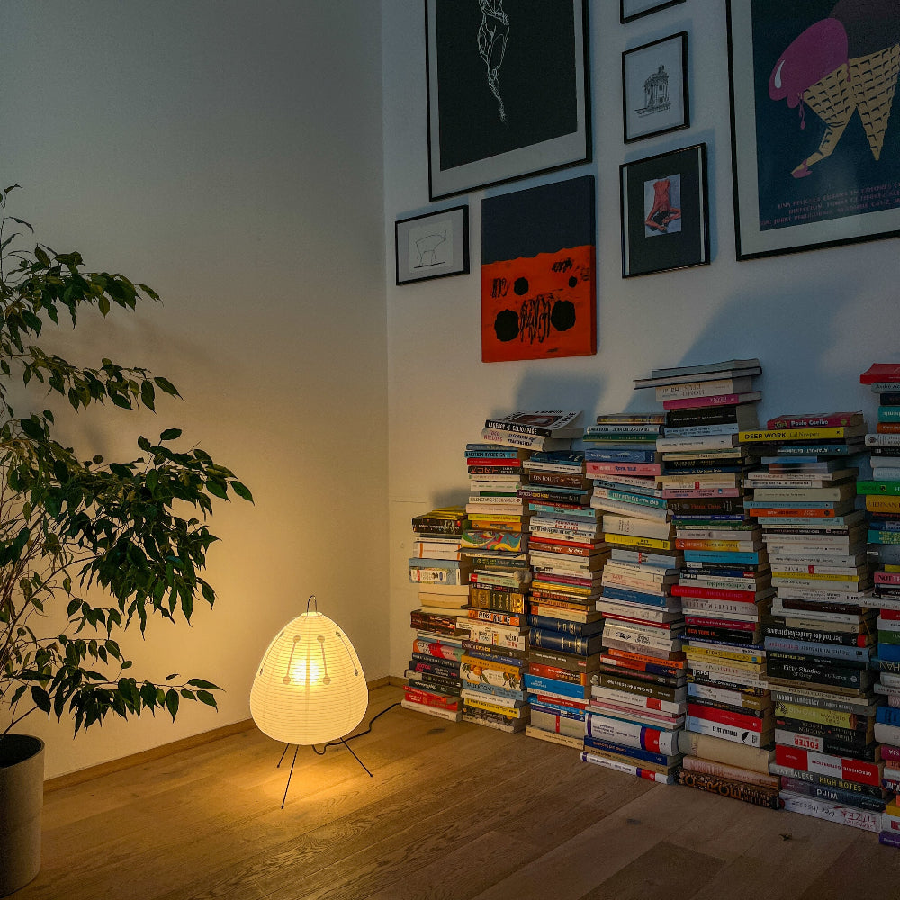 Diese minimalistische Papierlampe mit schwarzem Metallgestell sorgt für warmes Licht in einer modernen Wohnung. Sie steht auf einem hellen Holzboden neben einem hohen Bücherstapel und einer Pflanze.