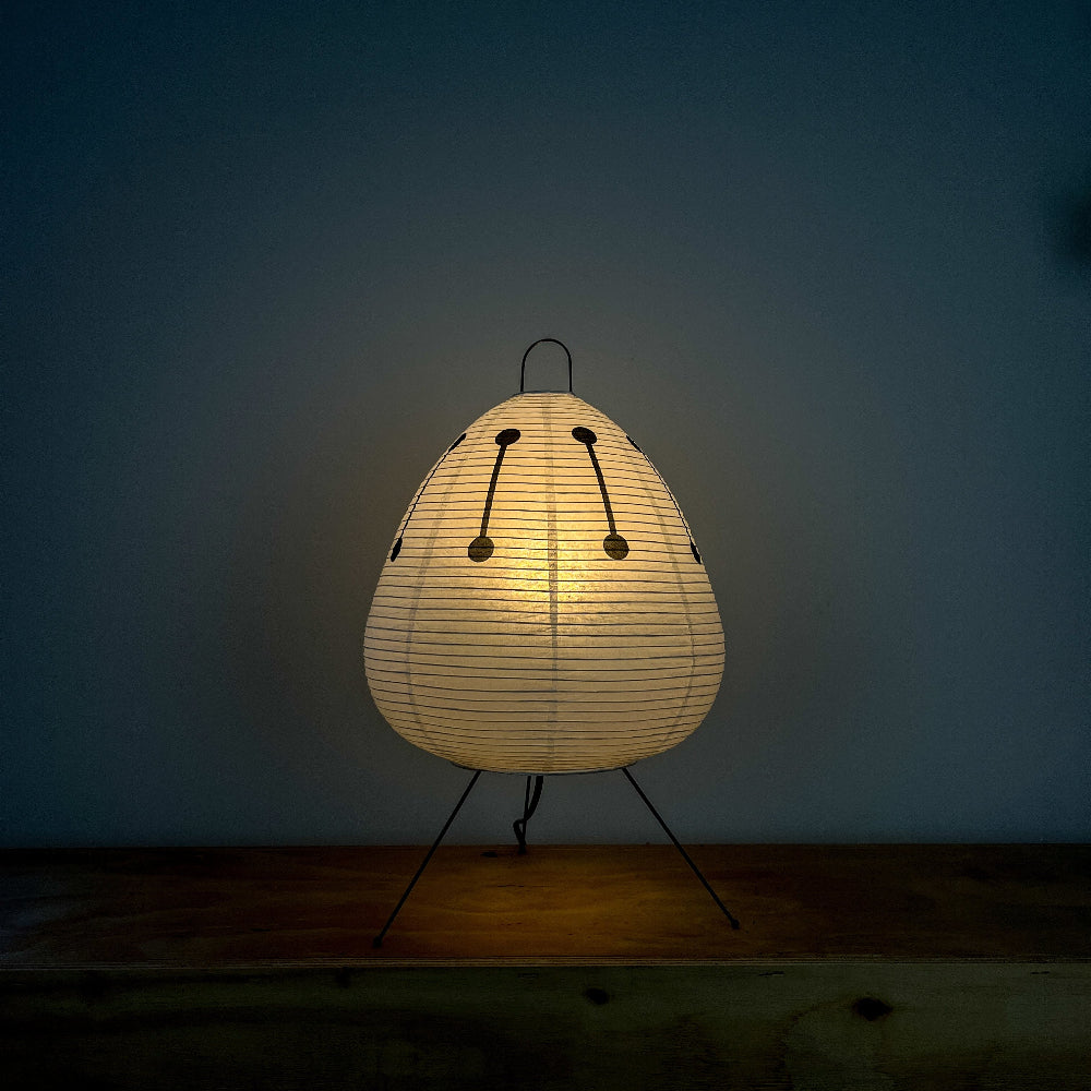 Die japanische Papierlampe mit schwarzem Muster strahlt warmes Licht aus. Sie steht auf einer Holzkommode vor einer dunkelblauen Wand. Ein gemütliches Ambiente für entspannte Lesestunden.