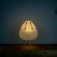 Die japanische Papierlampe mit schwarzem Muster strahlt warmes Licht aus. Sie steht auf einer Holzkommode vor einer dunkelblauen Wand. Ein gemütliches Ambiente für entspannte Lesestunden.