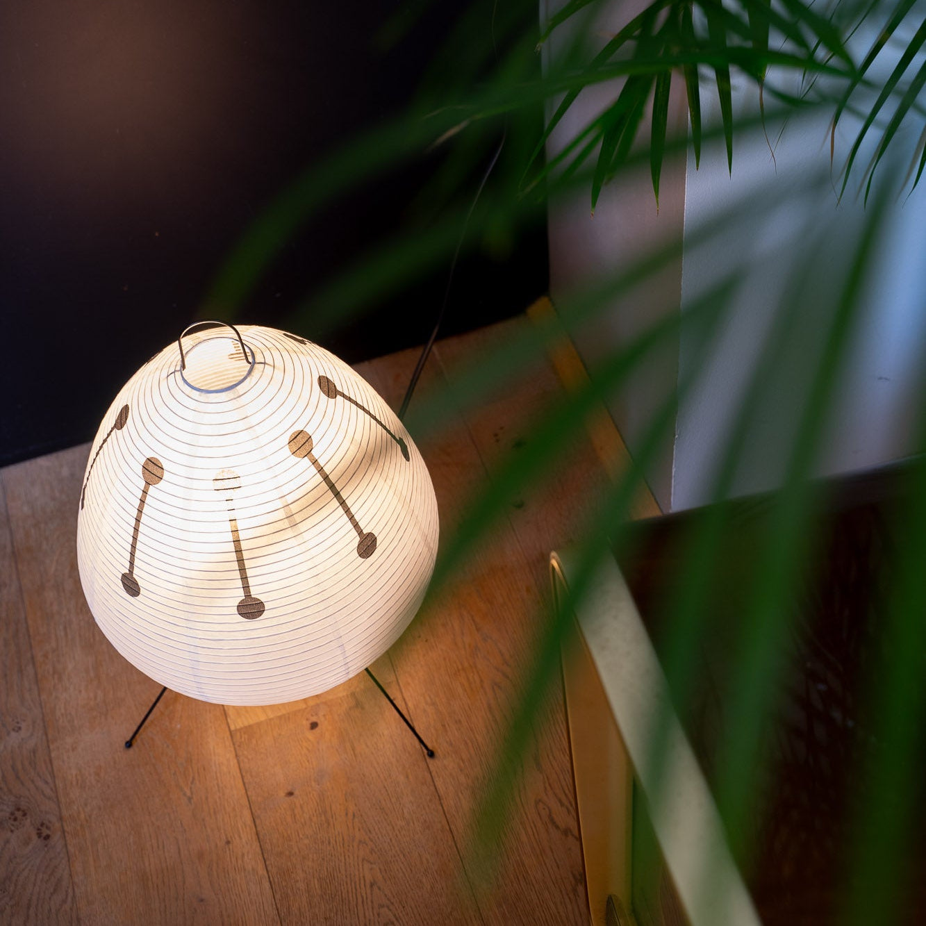 Die Papierlampe mit feinem schwarzem Muster und zarten Papierstreifen wird von oben fotografiert. Das warme Licht sorgt für eine angenehme Atmosphäre. Darunter ein moderner Holzfußboden mit dunklen Akzenten.
