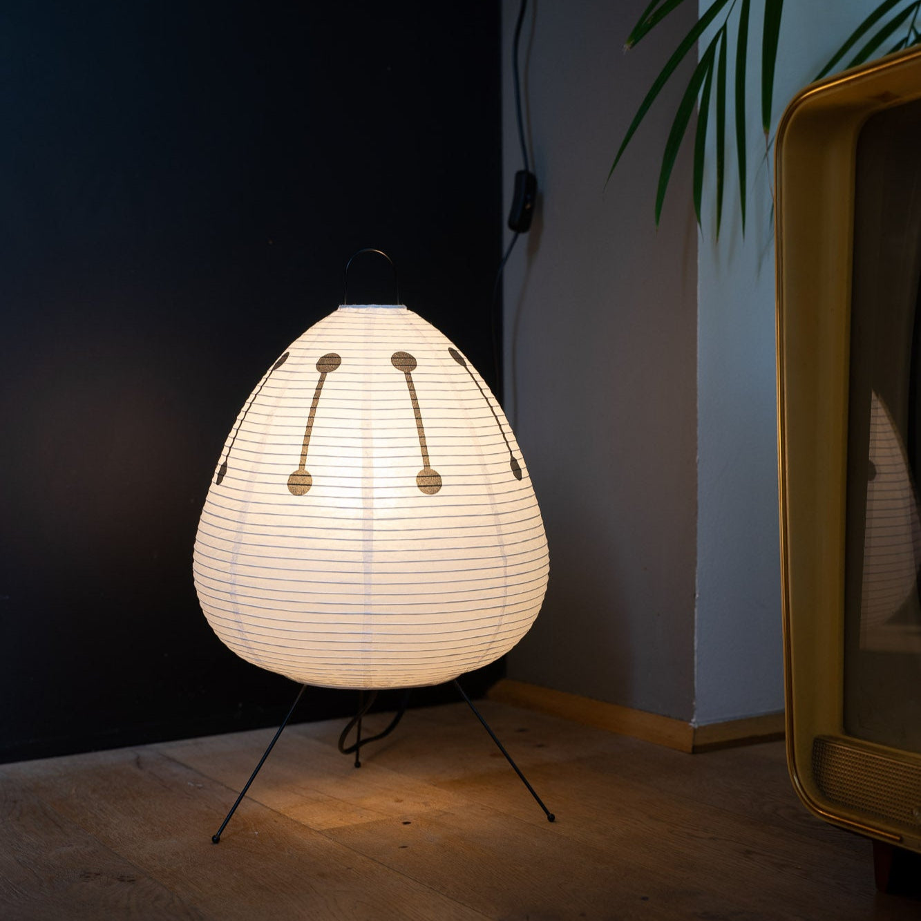 Die Papierlampe von GLOW UP STUDIO mit schwarzem Muster strahlt ein weiches Licht aus und steht in einer modernen Ecke mit dunkler Wand. Eine große Pflanze und ein Vintage-Radiomöbel ergänzen den Retro-Look.