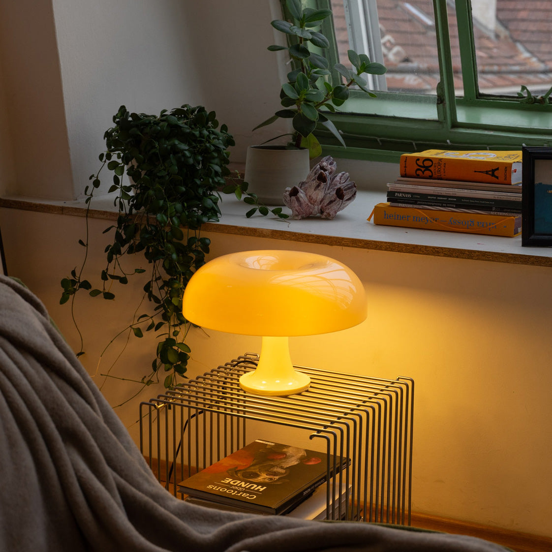 Gelbe Pilzlampe von GLOW UP STUDIO im Retro-Mushroom-Design auf einem modernen Gitter-Beistelltisch unter einem Fenster mit grüner Rahmenfarbe. Die Lampe spendet warmes, weiches Licht und schafft eine gemütliche Atmosphäre. Umgeben von Zimmerpflanzen, Kunstbüchern und einer beigen Decke auf dem Sofa, wird der Raum zum stylischen Rückzugsort mit Mid Century Flair und Space Age Charme.