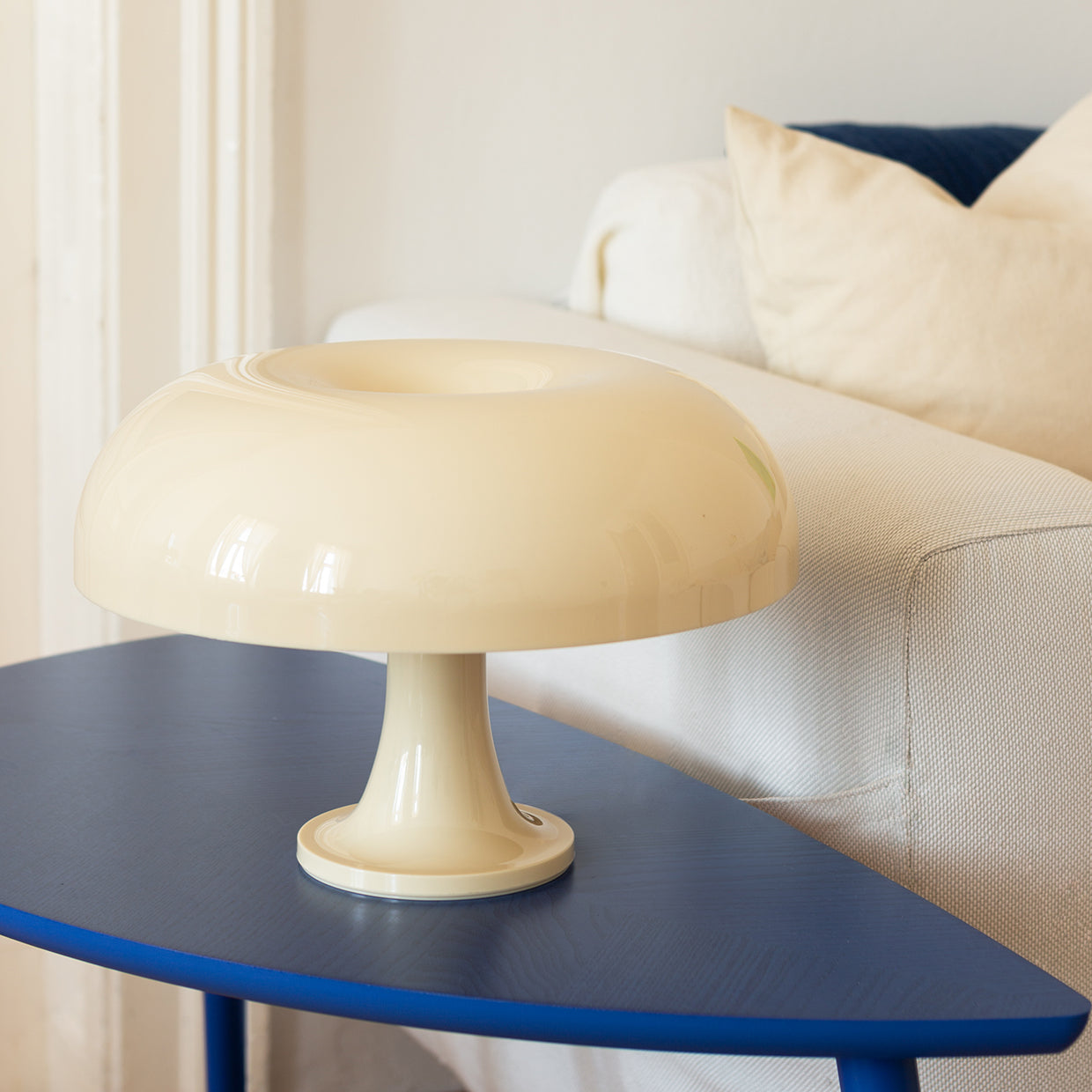 Die cremefarbene Vintage-Tischlampe steht auf einem blauen Design-Beistelltisch vor einem weißen Sofa mit beigen und dunkelblauen Kissen. Die weiche Beleuchtung und die Retro-Ästhetik schaffen eine stilvolle und einladende Atmosphäre.