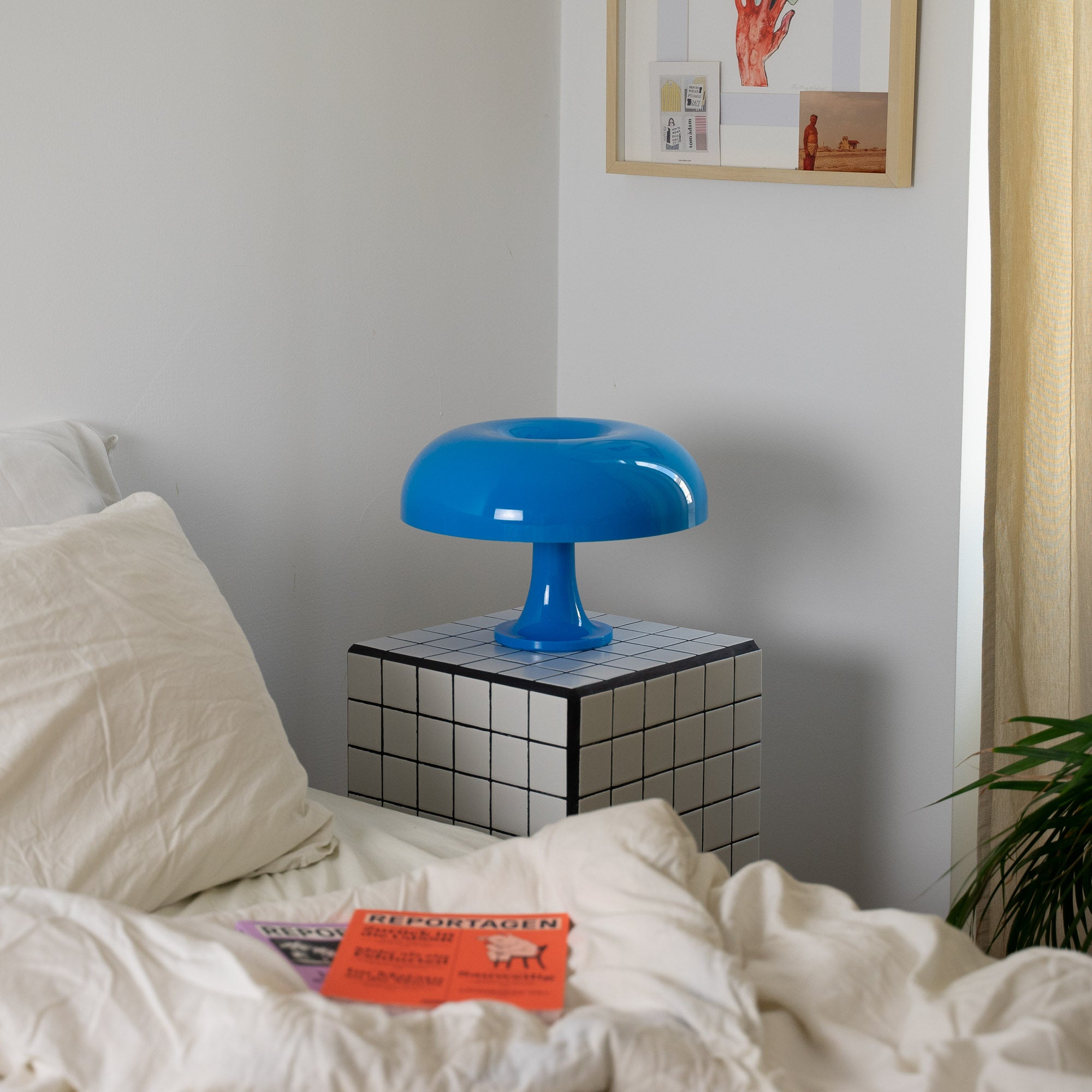 Die ikonische Retro-Lampe steht auf einem schwarz-weiß karierten Beistelltisch neben einem weißen Bett mit zerwühlter Decke. Wandbilder und eine helle Vorhangkulisse runden das moderne Interieur ab.