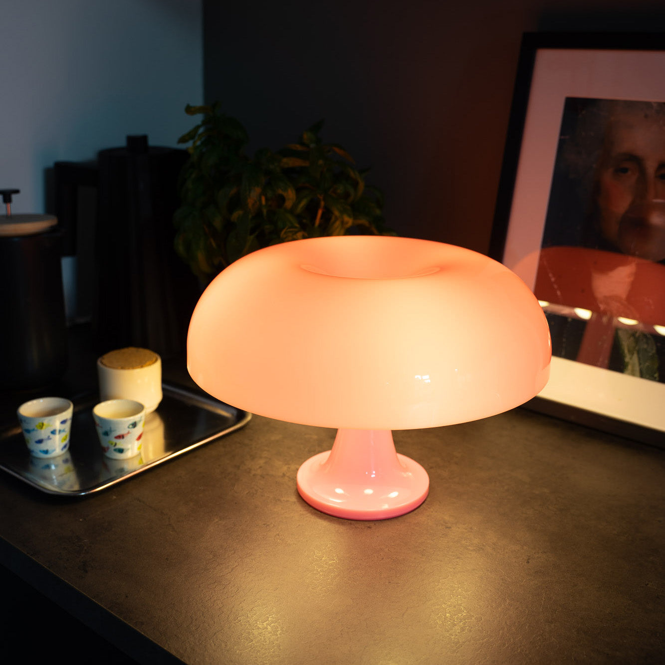 Leuchtende rosa Pilzlampe im Retro-Stil auf dunkler Arbeitsplatte, daneben Espressotassen, Pflanze und gerahmtes Porträt – stylische Mid-Century Tischlampe für gemütliches Vintage-Ambiente.