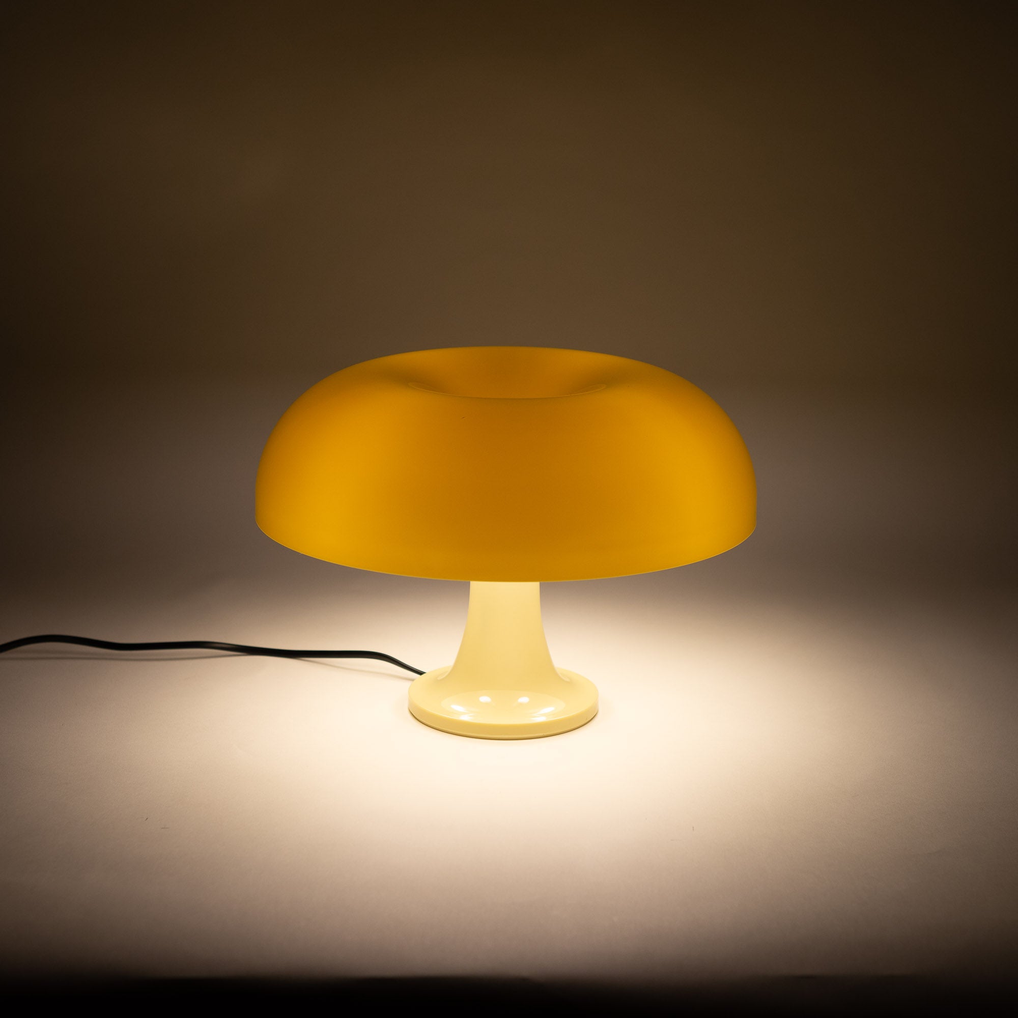 Die cremefarbene Pilzlampe mit glänzendem Acrylschirm und geschwungener Form steht auf einer weißen Oberfläche vor neutralem Hintergrund. Das hochwertige Material und die ikonische Form machen sie zu einem Designklassiker.