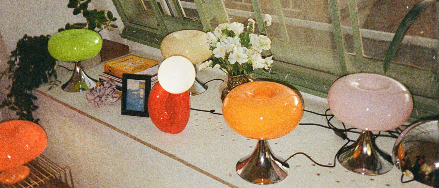Fensterbank mit bunten Midcentury-Tischlampen in Grün, Creme, Rot, Orange und Rosa auf verchromten Füßen. Dazwischen weiße Blumen in goldener Vase, Pflanzen, Bücher und ein kleines gerahmtes Bild sorgen für eine gemütliche Retro-Atmosphäre.