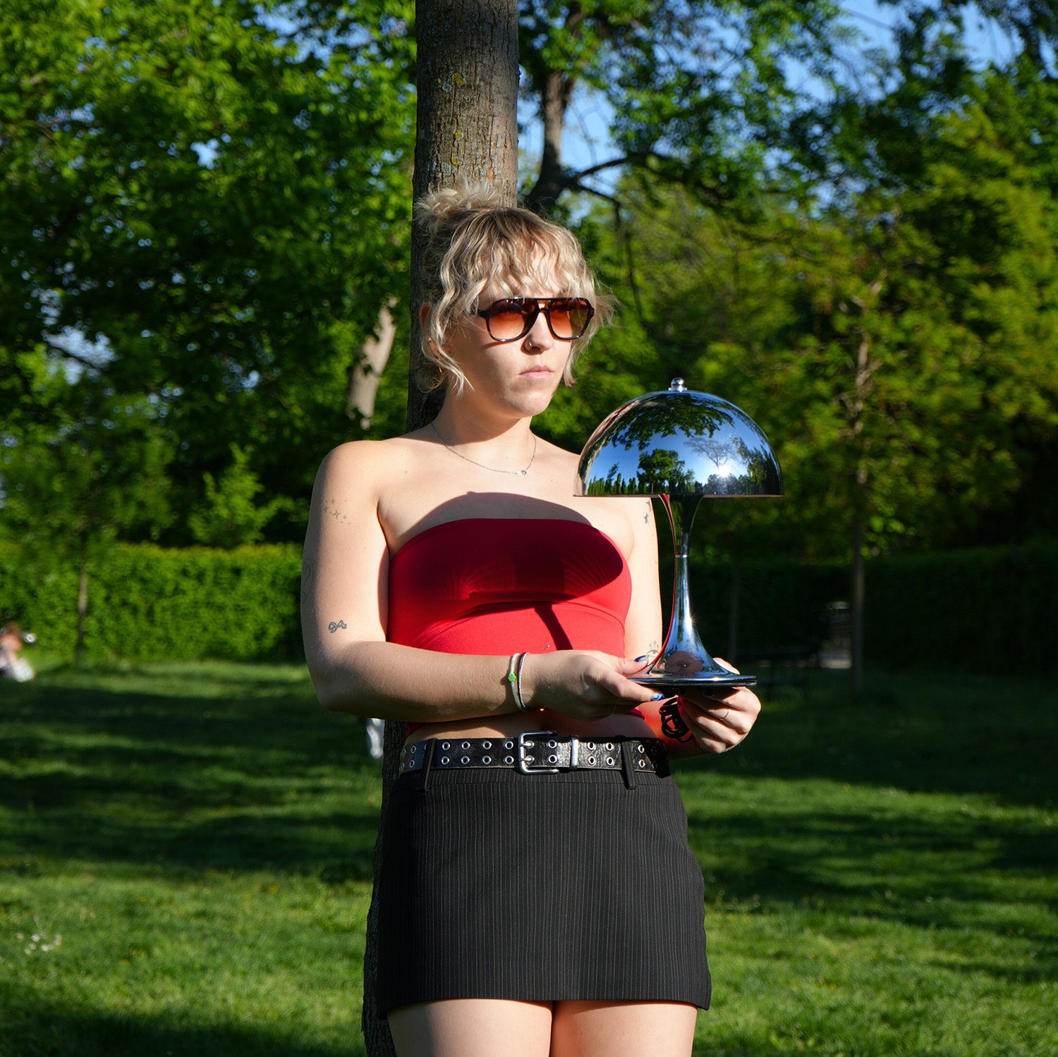 Retro Tischlampe Bell in Chrom von GLOW UP STUDIO – glänzende Designlampe im Mid Century Stil, outdoor im Park mit Person im roten Top.