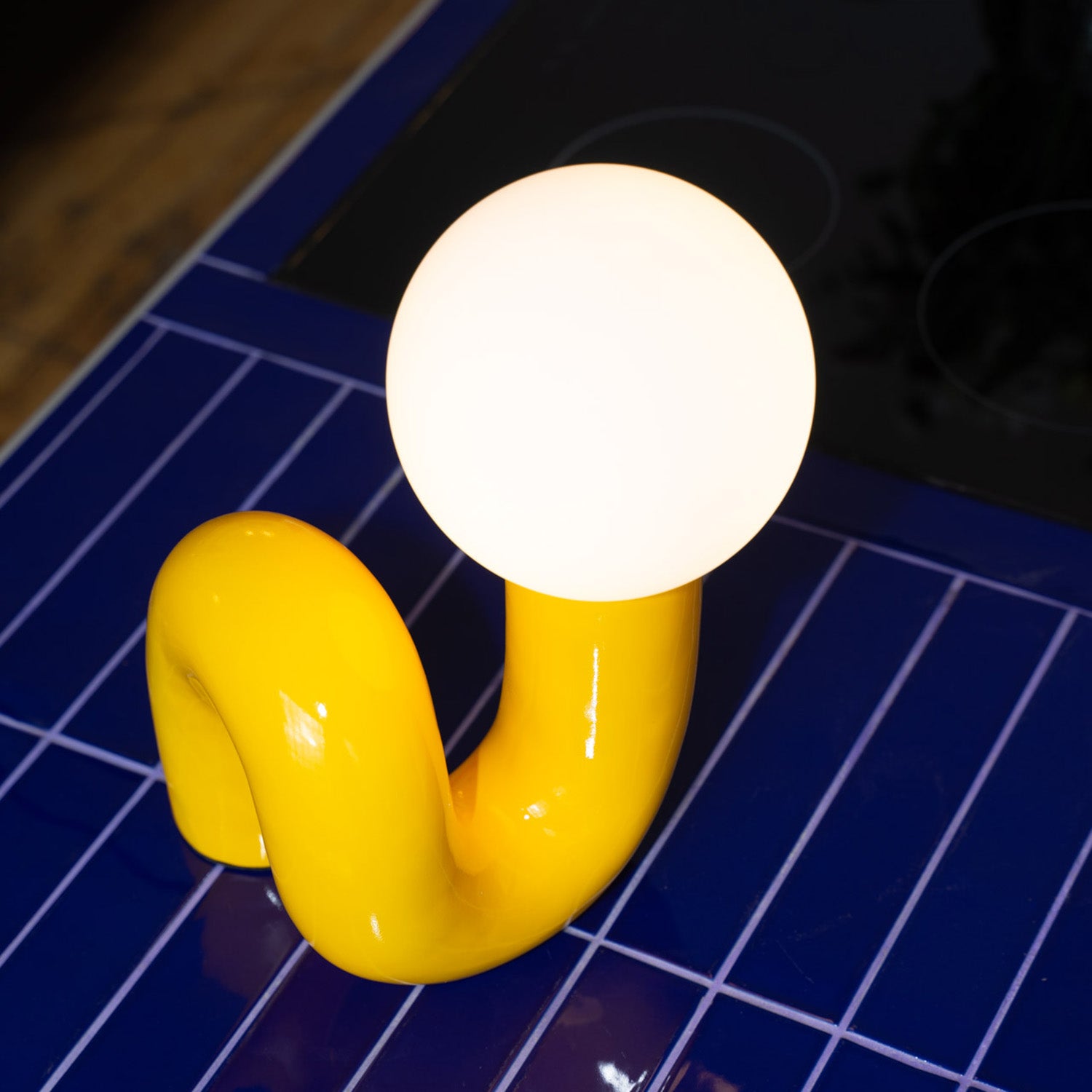 Retro Tischlampe Snake in Gelb von GLOW UP STUDIO – organische Designlampe mit Kugel-Leuchtkopf als Nahaufnahme auf blau gefliester Oberfläche.