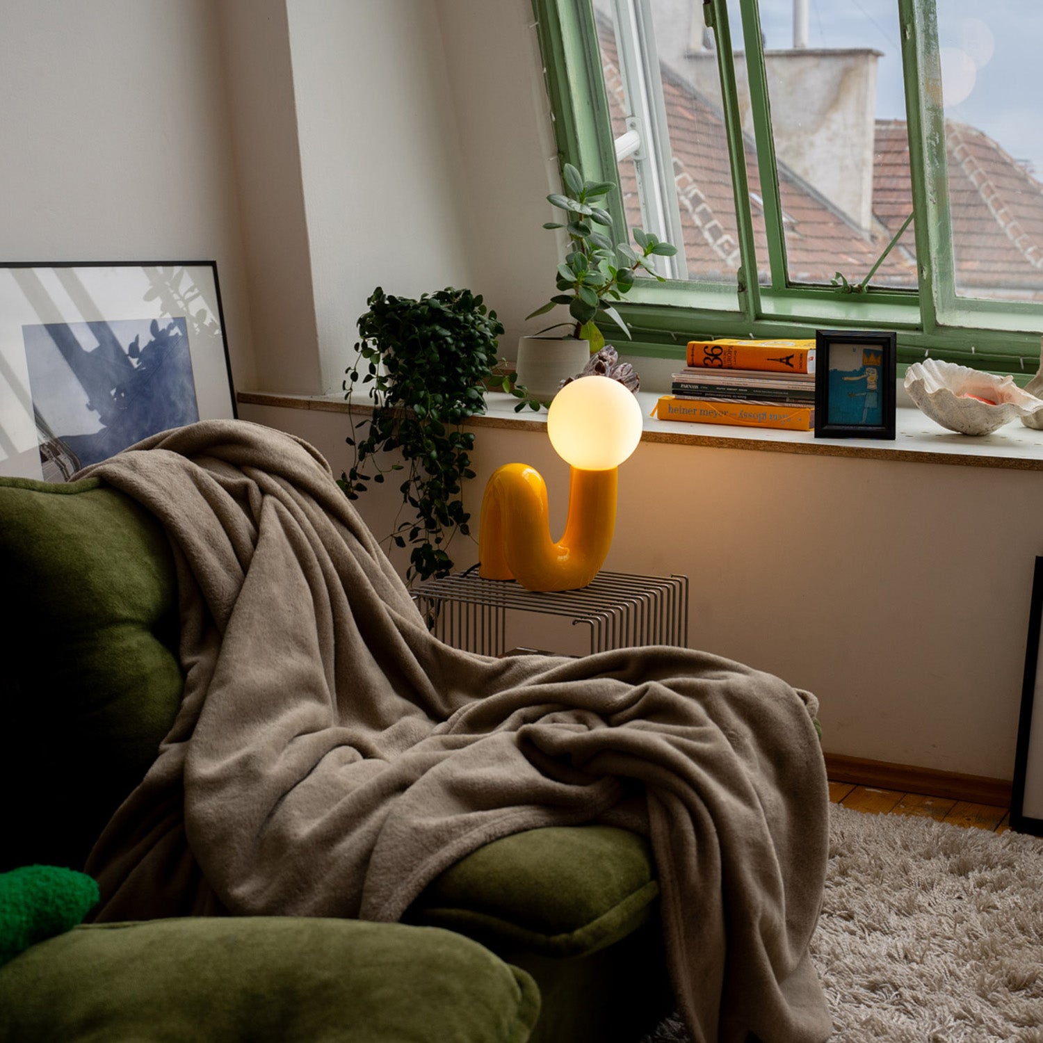 Retro Tischlampe Snake in Gelb von GLOW UP STUDIO – organische Lampe mit Kugel-Leuchtkopf auf Metalltisch vor Fensterbank, gemütliches Wohnzimmer mit Sofa, Pflanzen und Decke.