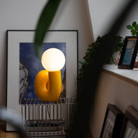 Retro Tischlampe Snake in Gelb von GLOW UP STUDIO – organische Designlampe mit Kugel-Leuchtkopf auf Metalltisch, gemütliches Wohnzimmer mit Kunst und Pflanzen.