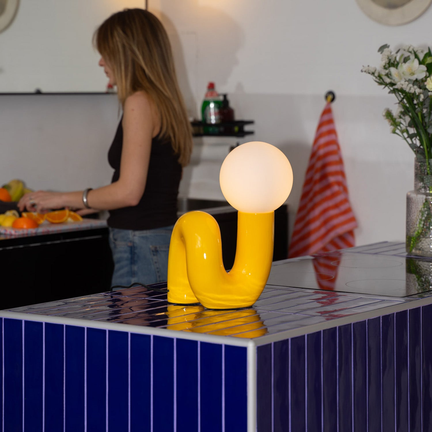 Retro Tischlampe Snake in Gelb von GLOW UP STUDIO – Designlampe mit Kugel-Leuchtkopf auf blau gefliester Kücheninsel, modernes Interior mit Frau im Hintergrund.