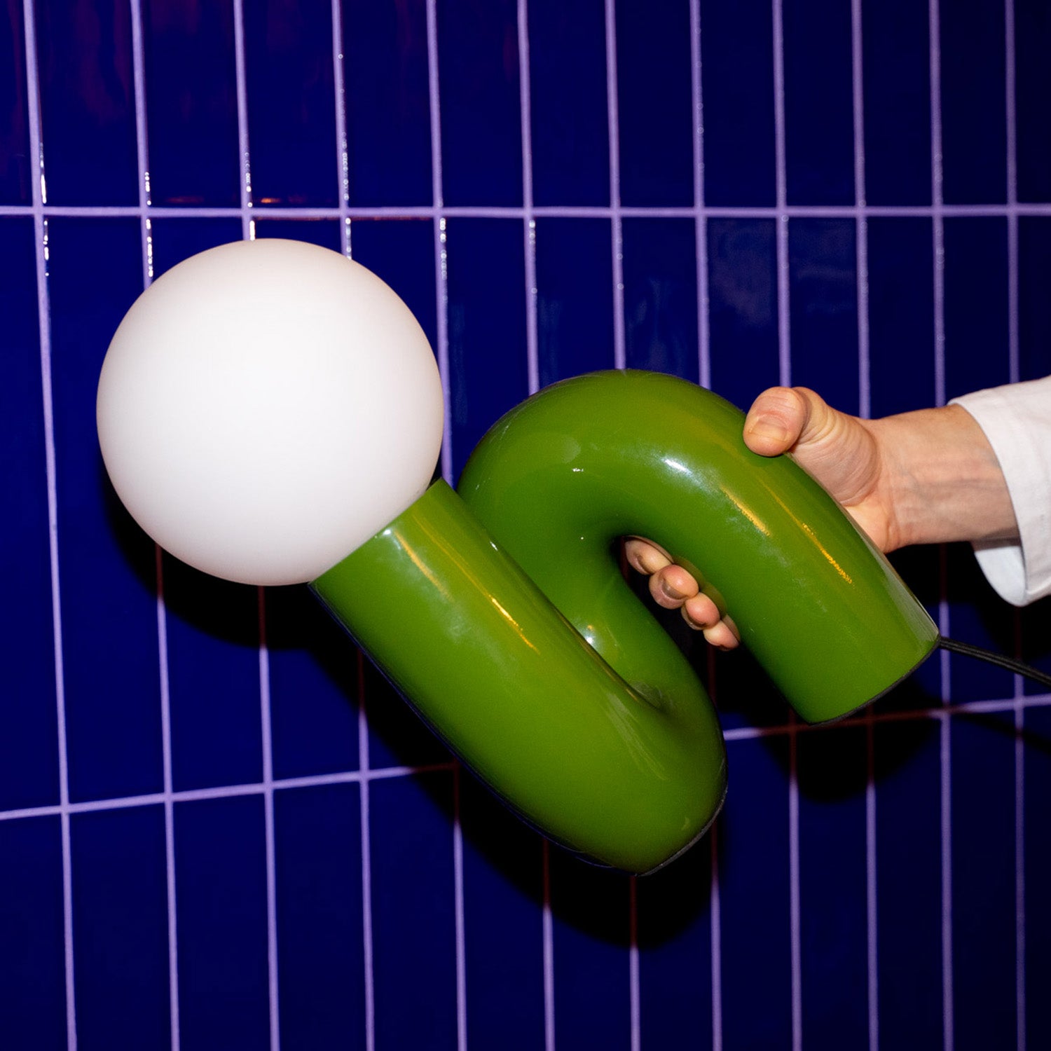 Retro Tischlampe Snake in Grün von GLOW UP STUDIO – organische Designlampe mit Kugel-Leuchtkopf, im Hand gehalten vor blau gekachelter Wand.
