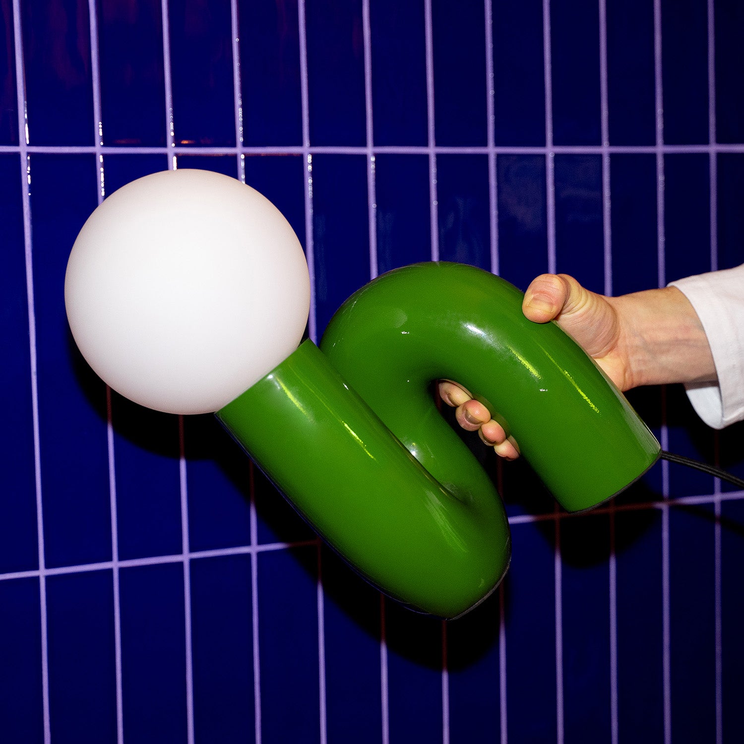 Retro Tischlampe Snake in Grün von GLOW UP STUDIO – organische Designlampe mit Kugel Leuchtkopf, im Hand gehalten vor blau gekachelter Wand.