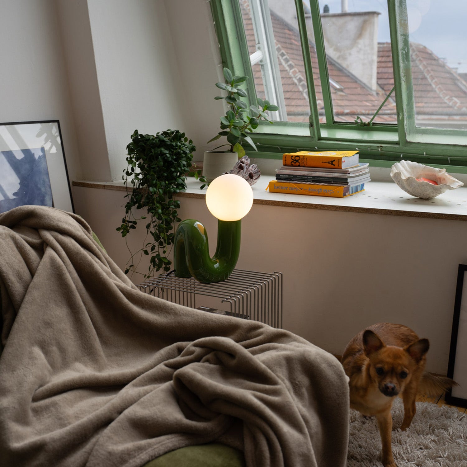 Retro Tischlampe Snake in Grün von GLOW UP STUDIO – Designlampe mit Kugel-Leuchtkopf auf Metalltisch vor Fensterbank mit Pflanzen und Büchern, gemütliches Wohnzimmer mit Hund.
