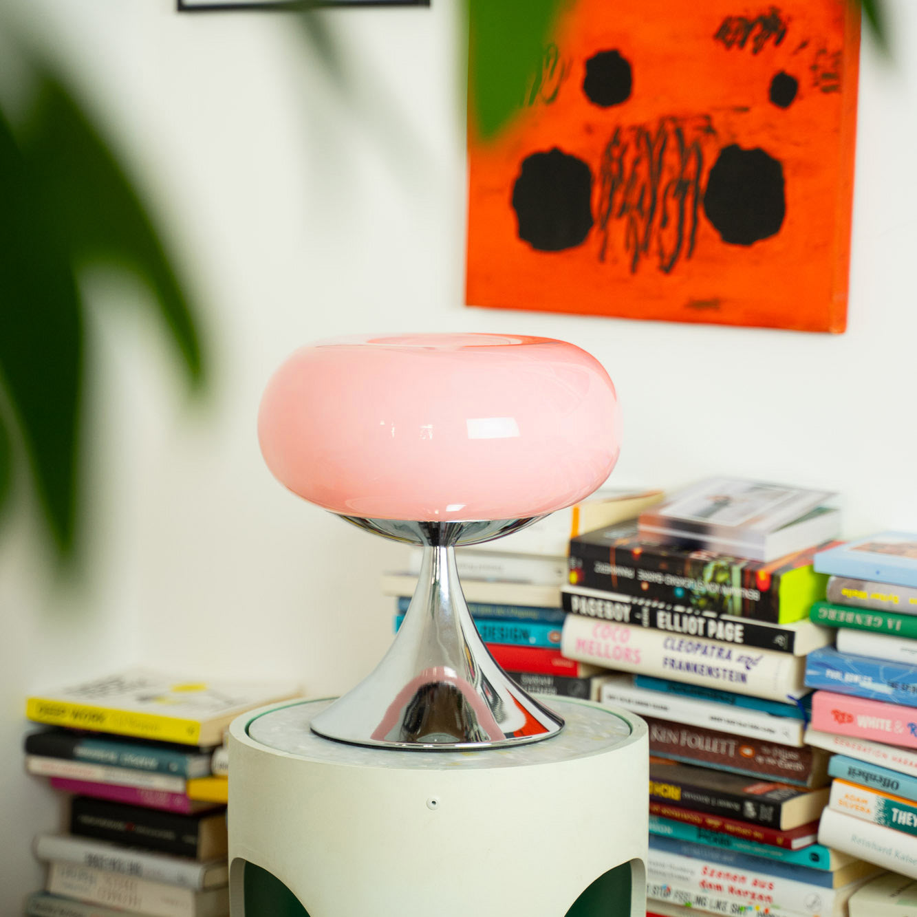Pastellrosa Space Age Tischlampe auf einem modernen Beistelltisch, umgeben von Büchern. Ein abstraktes Kunstwerk in Orange und Grün verleiht dem Hintergrund einen kreativen Akzent.