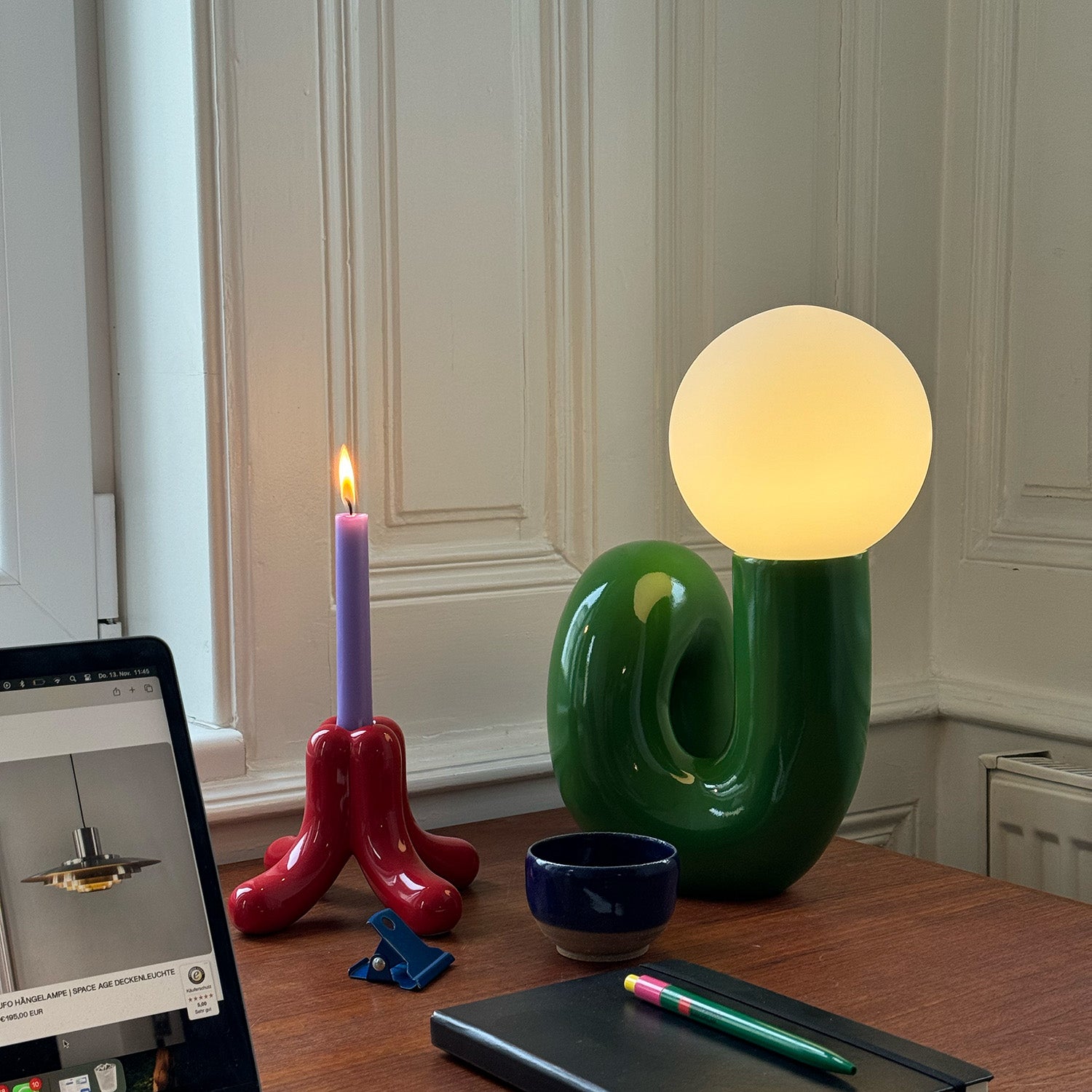 Grünes Snake Lampe von GLOW UP STUDIO im Retro Design auf einem Holztisch, daneben eine brennende lilafarbene Kerze im roten Kerzenhalter, eine kleine blaue Tasse, ein Notizbuch mit bunten Stiften und ein Laptop am Bildrand.