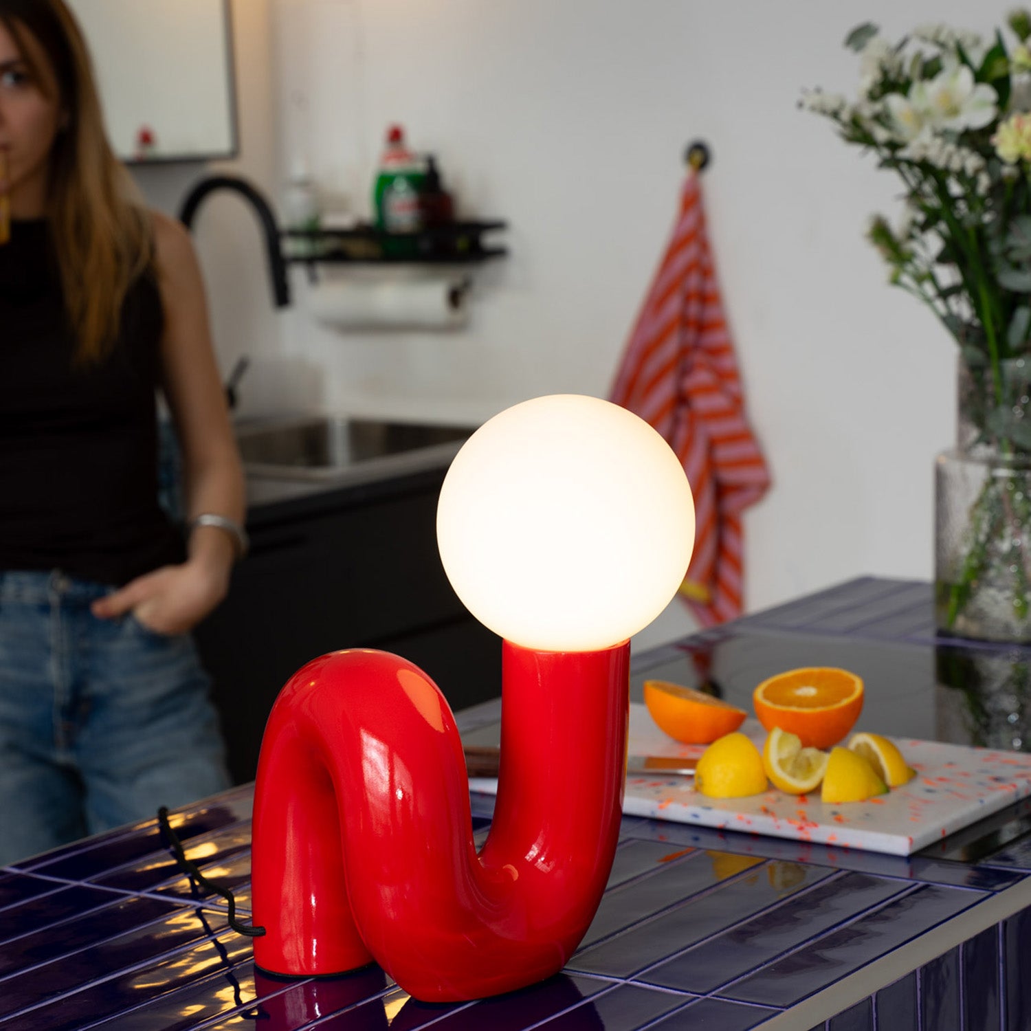 Rote Designlampe mit leuchtender Kugel auf dunkelblauer Kücheninsel mit Fliesen. Dahinter Zitrusfrüchte auf Schneidebrett, Vase mit weißen Blumen und gestreiftes Geschirrtuch. Im Hintergrund Frau in Jeans und dunklem Top an der schwarzen Spüle.