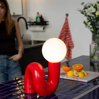 Rote Designlampe mit leuchtender Kugel auf dunkelblauer Kücheninsel mit Fliesen. Dahinter Zitrusfrüchte auf Schneidebrett, Vase mit weißen Blumen und gestreiftes Geschirrtuch. Im Hintergrund Frau in Jeans und dunklem Top an der schwarzen Spüle.