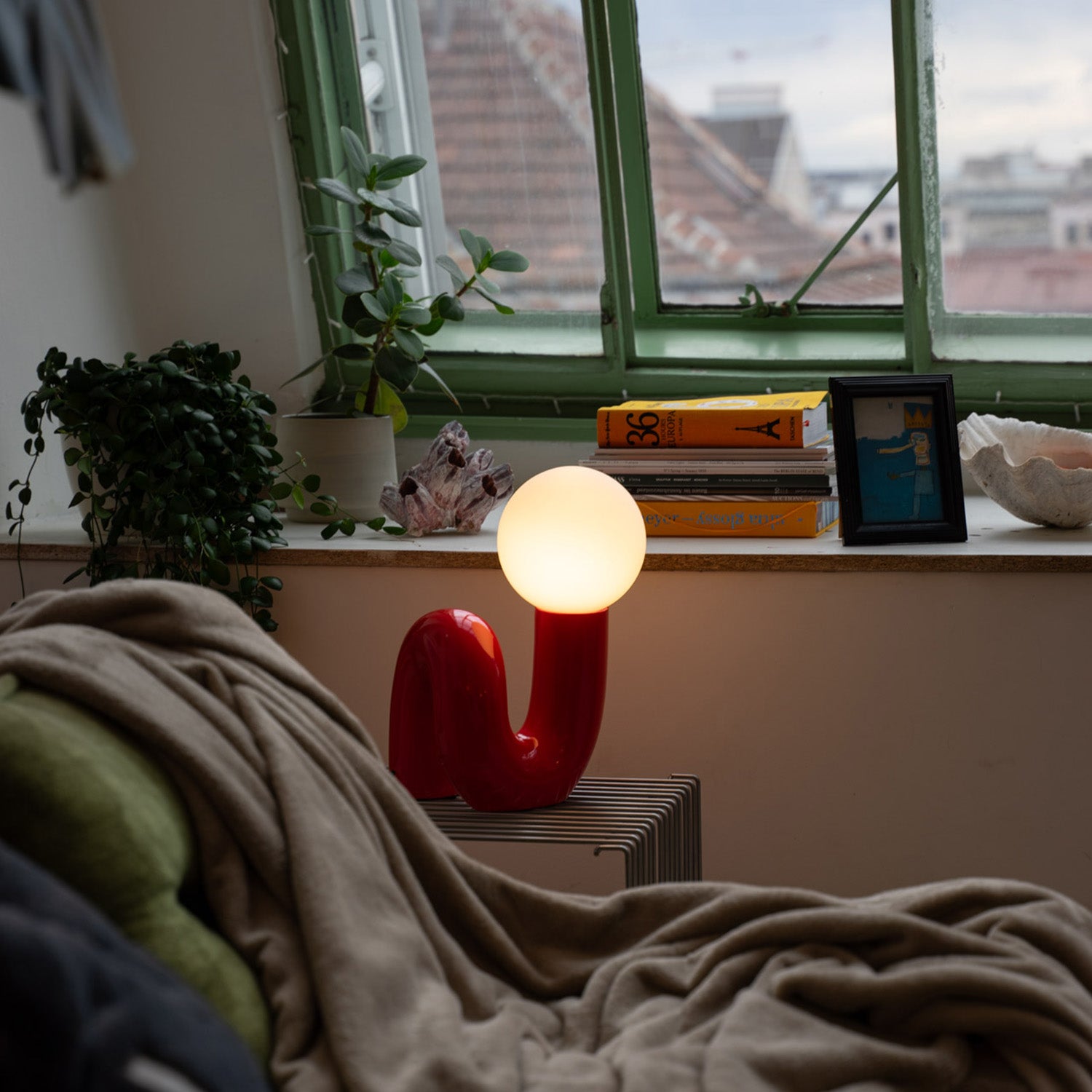 Rote Designlampe mit leuchtender Kugel auf Drahttisch neben grünem Sessel mit beiger Decke. Auf der Fensterbank Bücher, Pflanze, Muschel und gerahmtes Bild. Grünes Fenster mit Blick auf Dächer und die Stadt im Hintergrund.