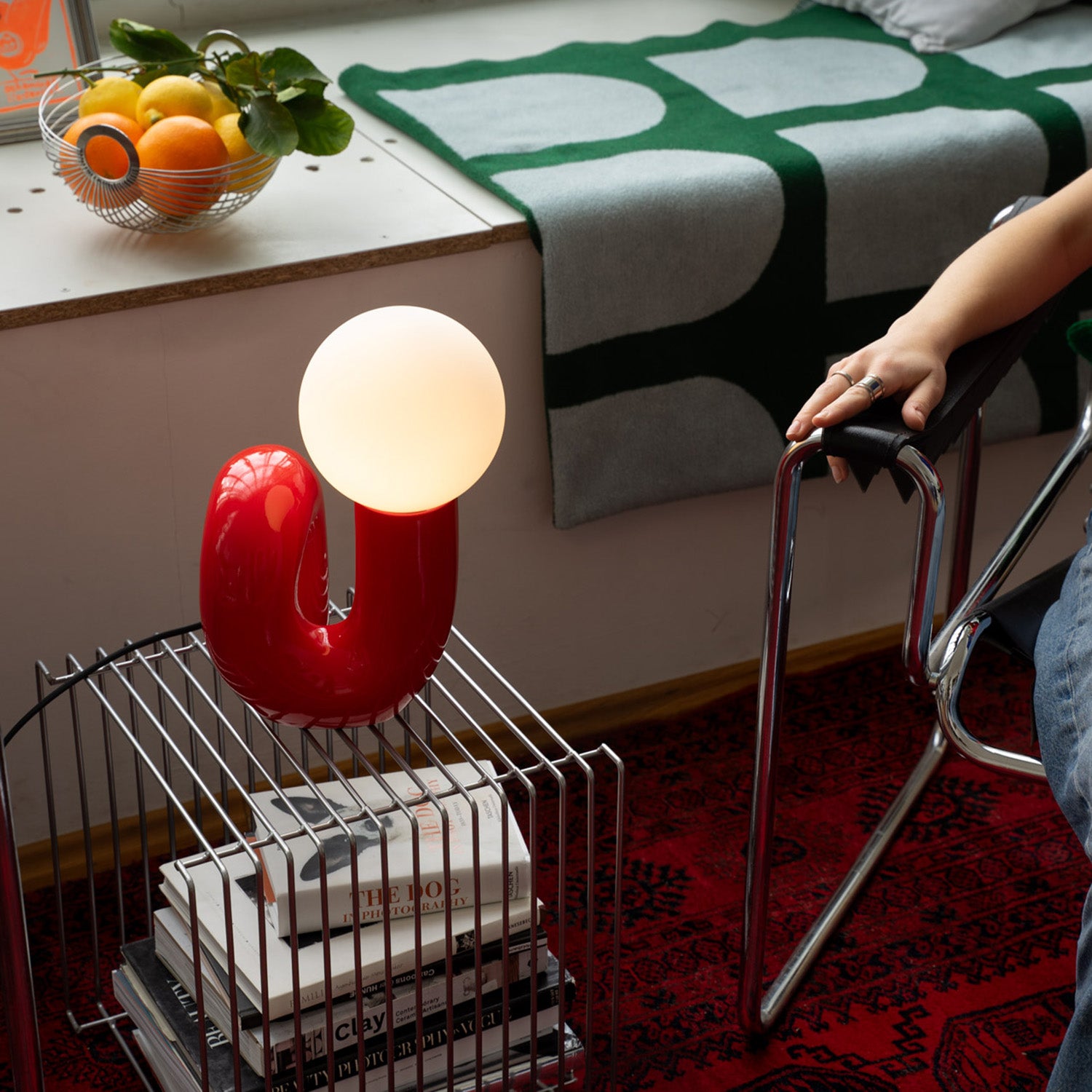 Rote Design-Tischlampe mit weißer Leuchtkugel auf Drahttisch mit Büchern, daneben ein verchromter Stuhl. Im Hintergrund Obstschale mit Zitrusfrüchten und grün-graue Decke auf Fensterbank, roter Teppich mit orientalischem Muster.