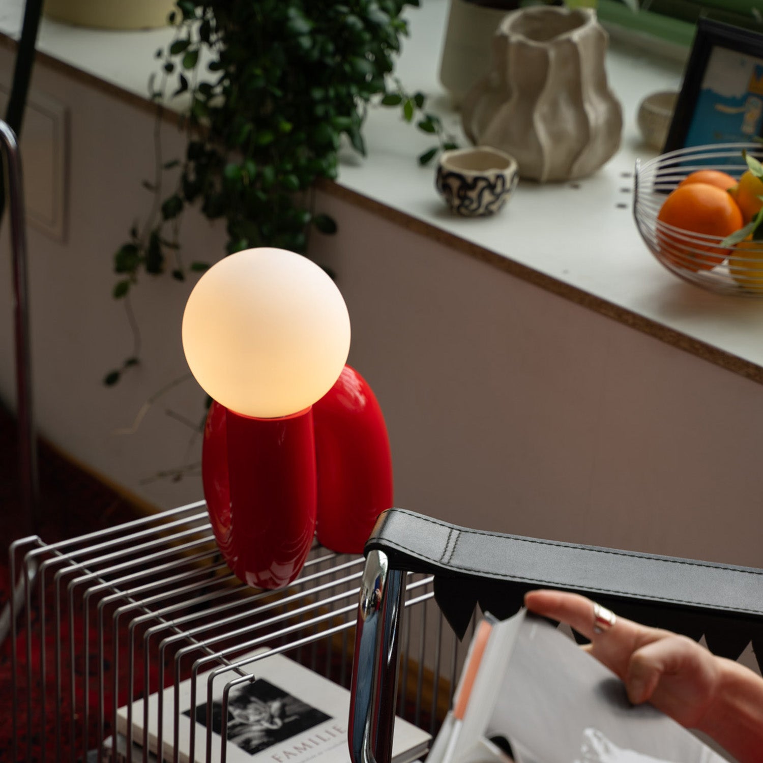 Rote Designlampe mit matter Leuchtkugel auf silbernem Drahttisch mit Büchern. Daneben verchromter Stuhl und eine Hand blättert in einem Buch. Auf der Fensterbank Obstschale, Keramik, Pflanze und hängende Efeuranke schaffen eine gemütliche Atmosphäre.