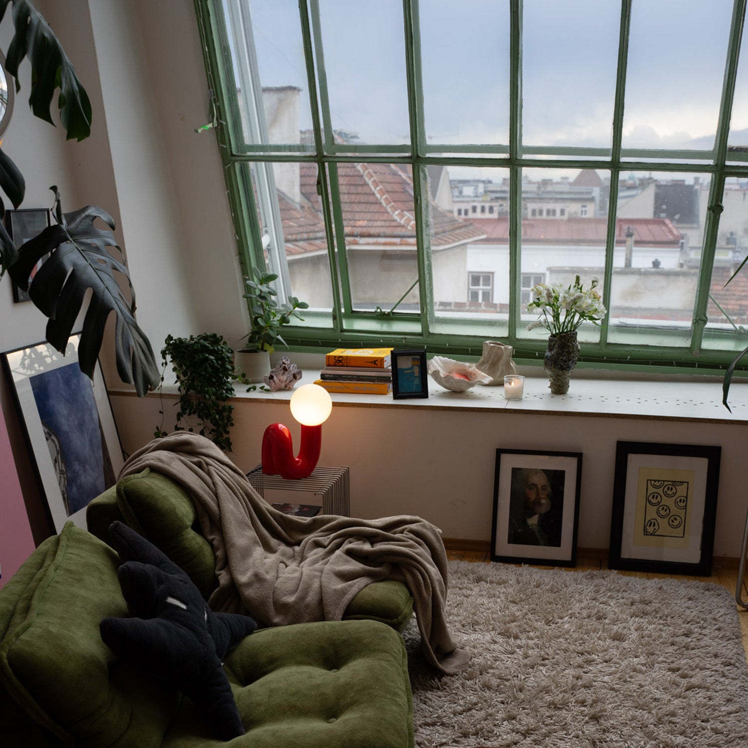 Gemütliche Wohnzimmerecke mit grünem Retro-Sessel, beiger Decke und schwarzem Kissen. Rote Designlampe mit Kugelleuchte auf Drahttisch, große Fensterfront mit Blick auf Dächer. Fensterbank mit Büchern, Pflanzen, Muschel und Vase mit weißen Blumen.