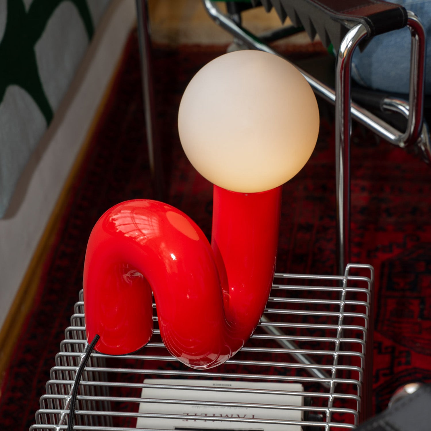 Rote Designlampe mit geschwungener Form und matter Leuchtkugel auf silbernem Drahttisch mit Büchern. Im Hintergrund verchromter Stuhl, roter Teppich mit Muster und grün-graue Decke an der Wand.