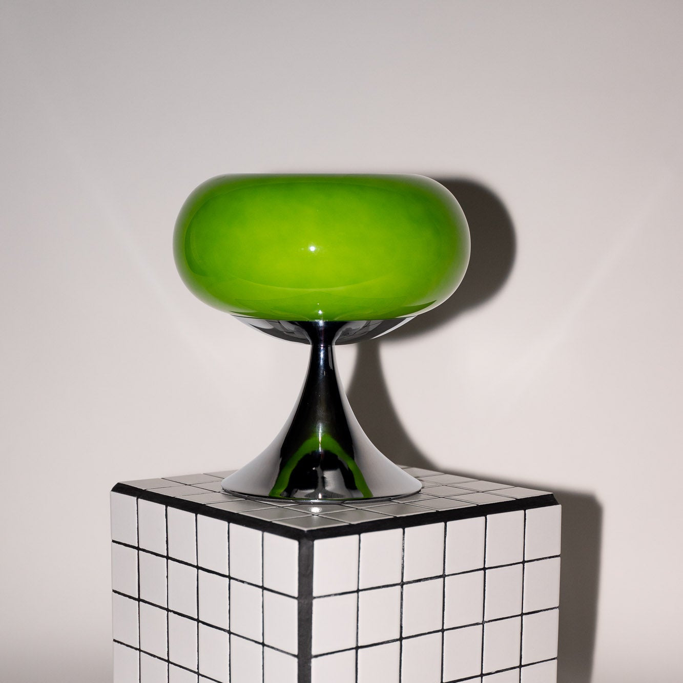 Kräftig grüne Glaslampe mit glänzendem Chromfuß auf einem grafischen Designwürfel. Der Kontrast zwischen dem satten Grün und dem minimalistischen Hintergrund betont das Space Age Design.