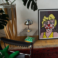 Verchromte Midcentury-Tischlampe auf silbernem Drahttisch mit Büchern, daneben bunter Pop-Art-Druck einer Frau mit gelbem Kleid. Schwarzer Lederstuhl mit grünem Kissen, große Monstera und roter Teppich sorgen für kreative Retro-Atmosphäre.