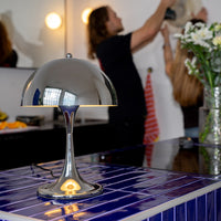Verchromte Midcentury-Tischlampe mit runder Kuppel auf dunkelblauer gefliester Kücheninsel. Im Hintergrund zwei Personen beim Dekorieren, weiße Blumen in Vase und gestreiftes Küchentuch sorgen für eine wohnliche Atmosphäre.