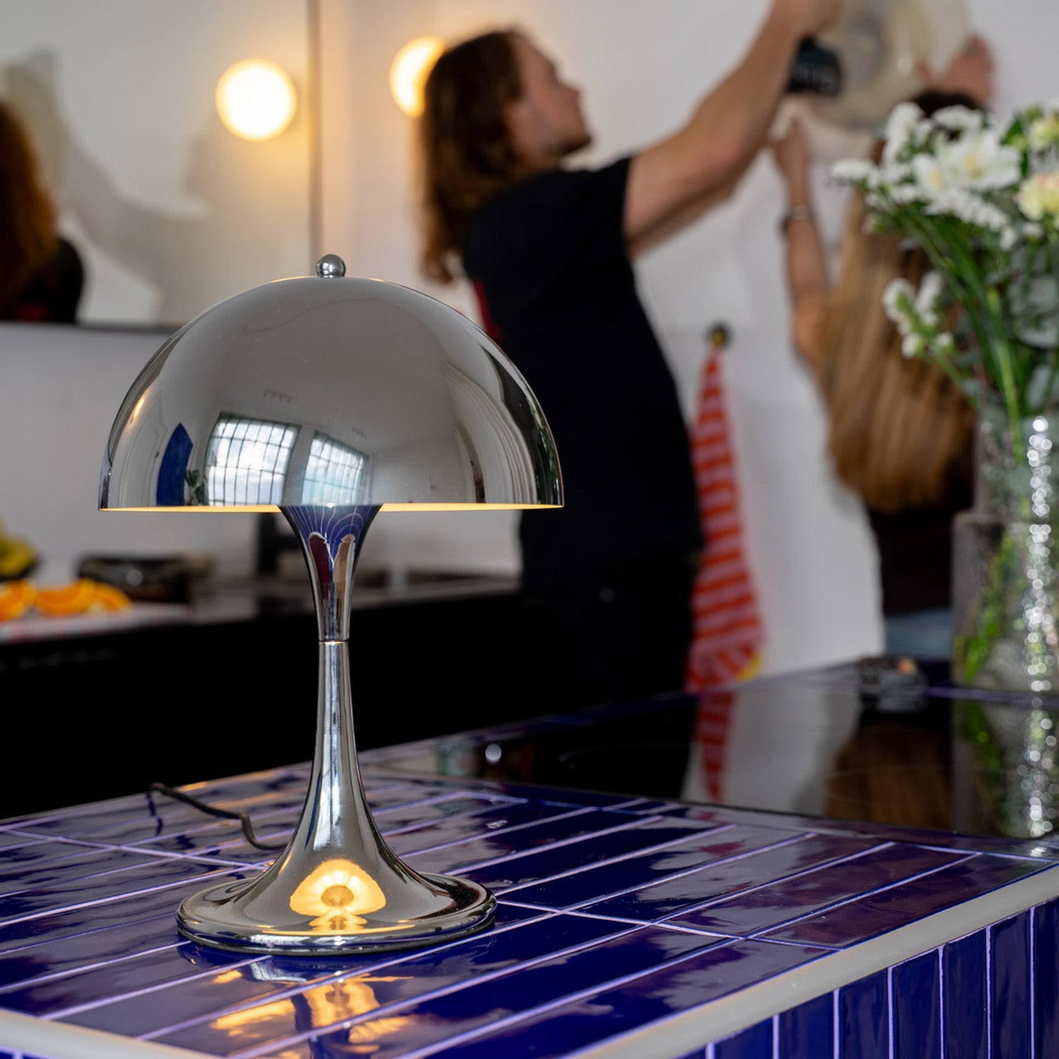 Verchromte Midcentury-Tischlampe mit runder Kuppel auf dunkelblauer gefliester Kücheninsel. Im Hintergrund zwei Personen beim Dekorieren, weiße Blumen in Vase und gestreiftes Küchentuch sorgen für eine wohnliche Atmosphäre.