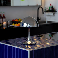 Verchromte Midcentury-Tischlampe mit halbrundem Schirm auf dunkelblauer gefliester Kücheninsel. Im Hintergrund schwarze Küche mit Obstschale, Kerze und Spüle, moderne Armaturen und aufgeräumte, minimalistische Atmosphäre.