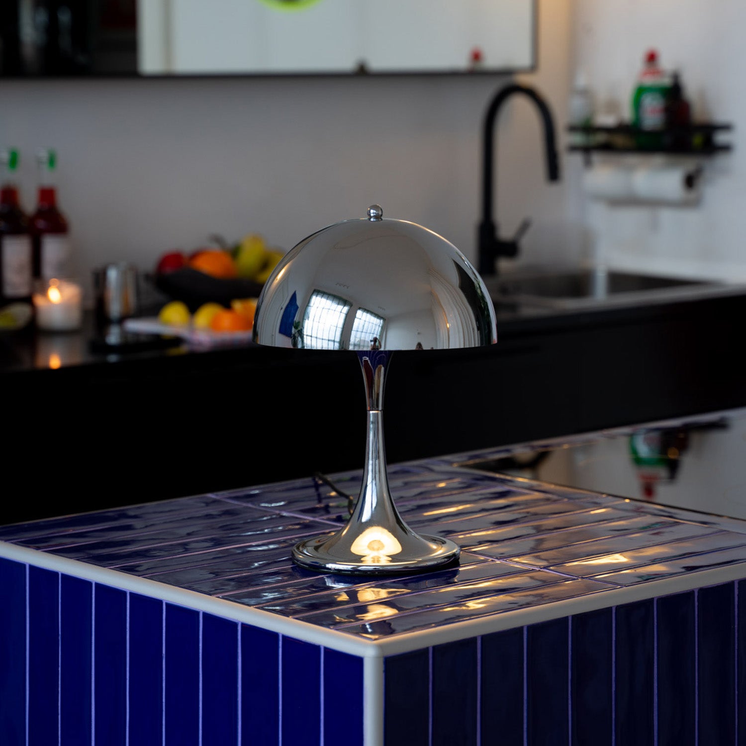 Verchromte Midcentury-Tischlampe mit halbrundem Schirm auf dunkelblauer gefliester Kücheninsel. Im Hintergrund schwarze Küche mit Obstschale, Kerze und Spüle, moderne Armaturen und aufgeräumte, minimalistische Atmosphäre.