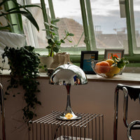 Verchromte Midcentury-Tischlampe auf silbernem Drahttisch vor einem großen Fenster mit grünem Rahmen. Auf der Fensterbank Pflanzen, Keramik, gerahmte Bilder und eine Obstschale mit Zitrusfrüchten sorgen für eine gemütliche Atmosphäre.