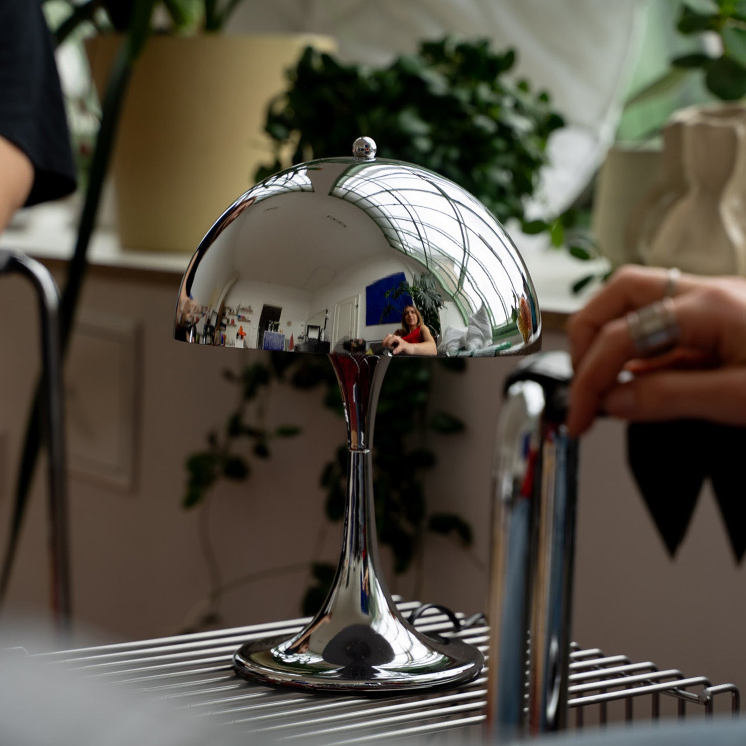 Verchromte Midcentury-Tischlampe mit glänzender Kuppel auf silbernem Drahttisch. Die Lampe spiegelt den Raum, eine Frau und große Fensterfront. Im Hintergrund grüne Pflanzen, Keramikvase und eine Hand am verchromten Stuhl.