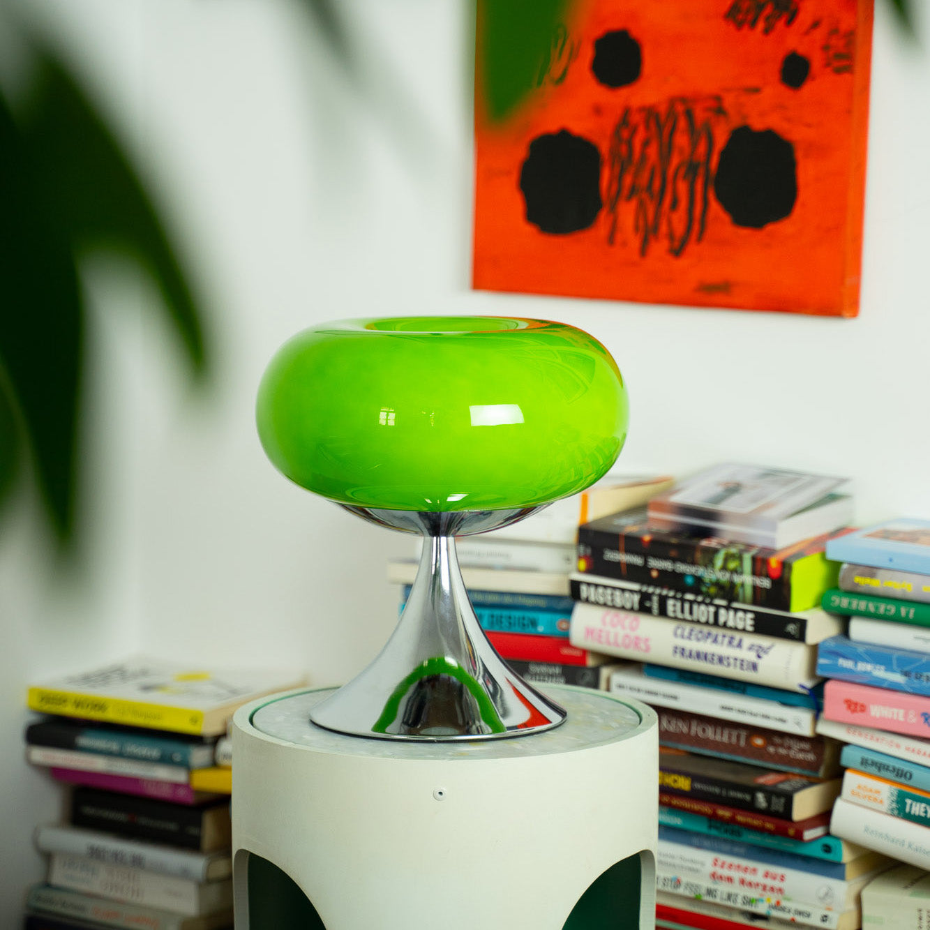 Die grüne Space Age Leuchte steht auf einem modernen Beistelltisch vor einem Bücherstapel. Ein leuchtend oranges Kunstwerk setzt einen starken Kontrast, während sanfte Schatten von einer Pflanze fallen.