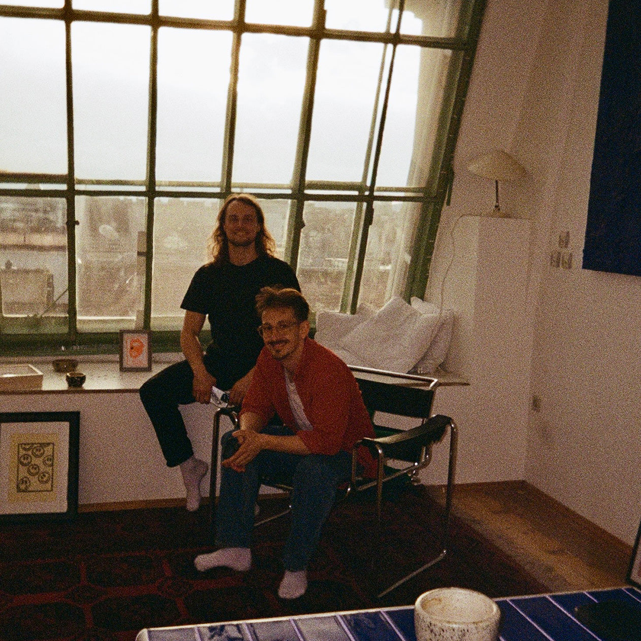 Zwei Männer in gemütlichem Wohnzimmer mit großer Fensterfront und Blick auf die Stadt. Einer sitzt auf einem Stuhl, der andere auf der Fensterbank. Roter Teppich, moderne Möbel, gerahmte Bilder und Kissen sorgen für eine wohnliche Atmosphäre.