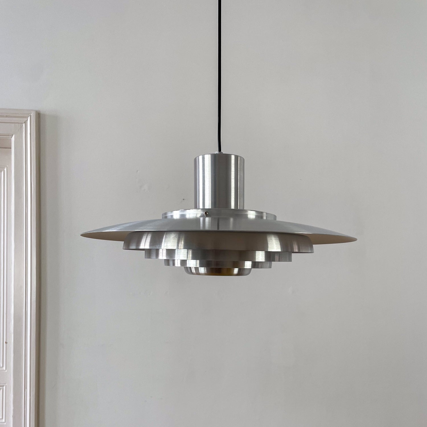 Gebürstete Chromlampe mit gestuftem Reflektorschirm, ideal für Esszimmer oder Büros. Der Hintergrund mit weißen Wänden und einer klassischen Altbau-Tür unterstreicht das reduzierte Design.