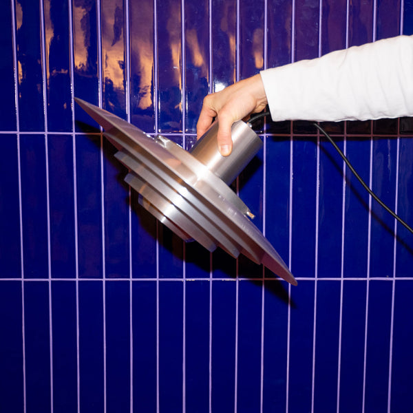 Silberne Midcentury-Pendelleuchte mit gestufter Metallstruktur wird von einer Hand vor glänzender, dunkelblauer Fliesenwand gehalten. Die geometrische Lampe setzt einen modernen, industriellen Akzent.