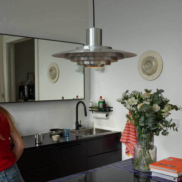 Moderne Küche mit schwarzer Arbeitsfläche, Spüle und großer Metall-Pendelleuchte im Midcentury-Stil. Auf der blauen Kochinsel ein Glasstrauß mit weißen Blumen und Kochbüchern, im Hintergrund Spiegel, Geschirrtuch und Spülutensilien.