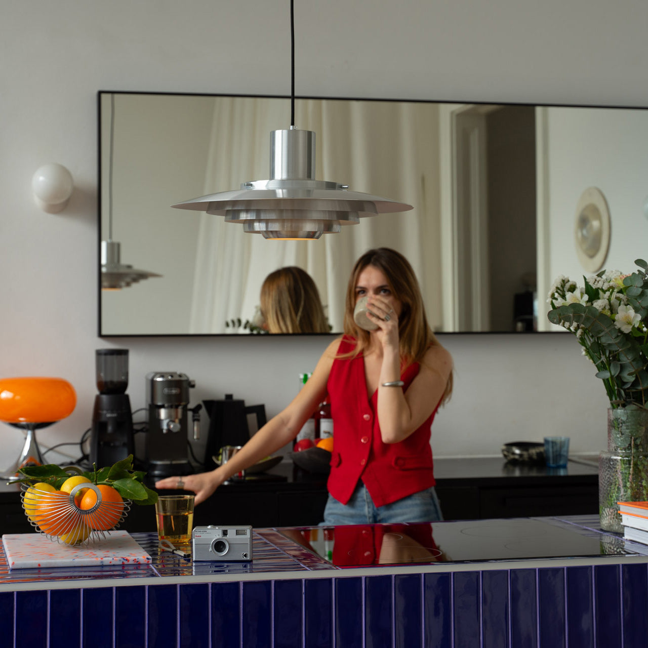 Moderne Küche mit blauer Flieseninsel und Metall-Pendelleuchte im Midcentury-Stil. Frau im roten Top trinkt aus einer Tasse. Auf der Insel Obstschale, Kamera und Blumenstrauß, im Hintergrund Espressomaschine, orange Tischlampe und Spiegelwand.