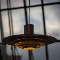 Elegante Midcentury-Pendelleuchte aus gebürstetem Metall mit mehreren abgestuften Ringen und warmem Licht. Im Hintergrund große Fensterfront mit Blick auf den Himmel bei Abenddämmerung.