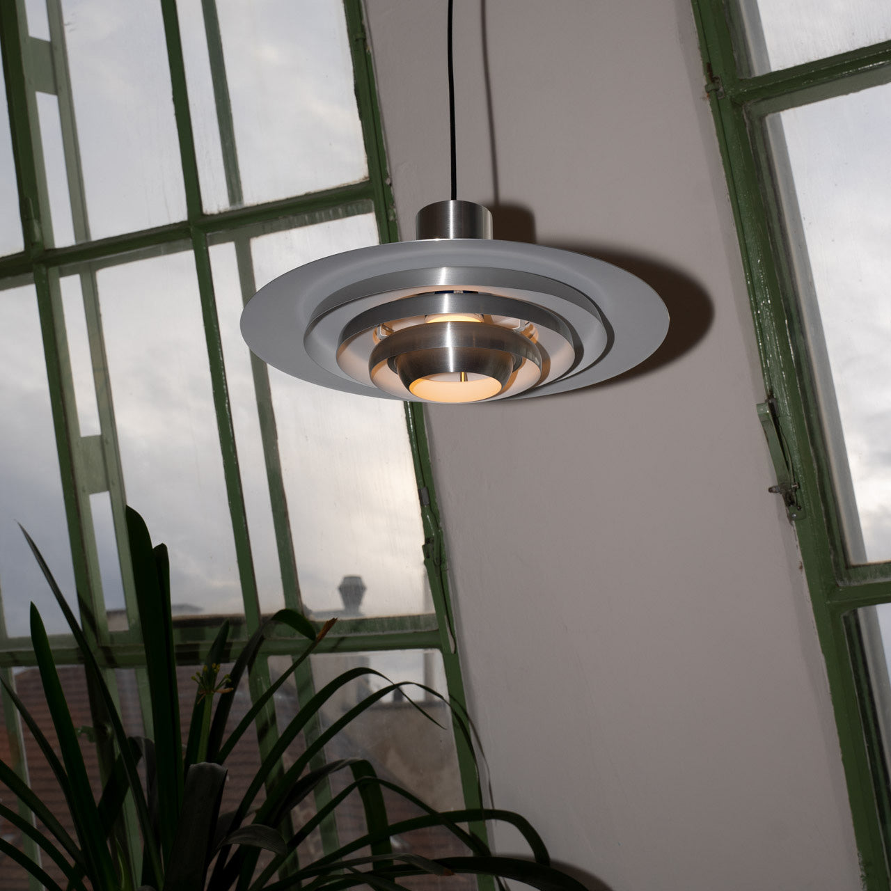 Moderne Midcentury-Pendelleuchte aus gebürstetem Metall mit runder, abgestufter Form und warmem Licht. Im Hintergrund große grüne Fensterfront mit Blick auf Dächer und Himmel, unten eine grüne Zimmerpflanze.