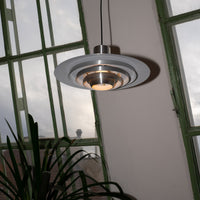 Moderne Midcentury-Pendelleuchte aus gebürstetem Metall mit runder, abgestufter Form und warmem Licht. Im Hintergrund große grüne Fensterfront mit Blick auf Dächer und Himmel, unten eine grüne Zimmerpflanze.