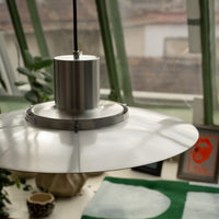 Nahaufnahme der Midcentury-Pendelleuchte Ufo Lampe aus Metall mit flachem, rundem Schirm. Im Hintergrund grüne Fensterrahmen, Pflanzen, Keramik, gerahmte Bilder und eine grün-gemusterte Decke auf der Fensterbank.
