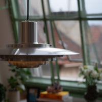 Silberne Midcentury-Pendelleuchte mit gestufter Metallform vor großer Fensterfront mit grünem Rahmen. Im Hintergrund Bücher, Pflanzen und ein Blumenstrauß auf der Fensterbank, Blick auf Dächer und den Himmel.