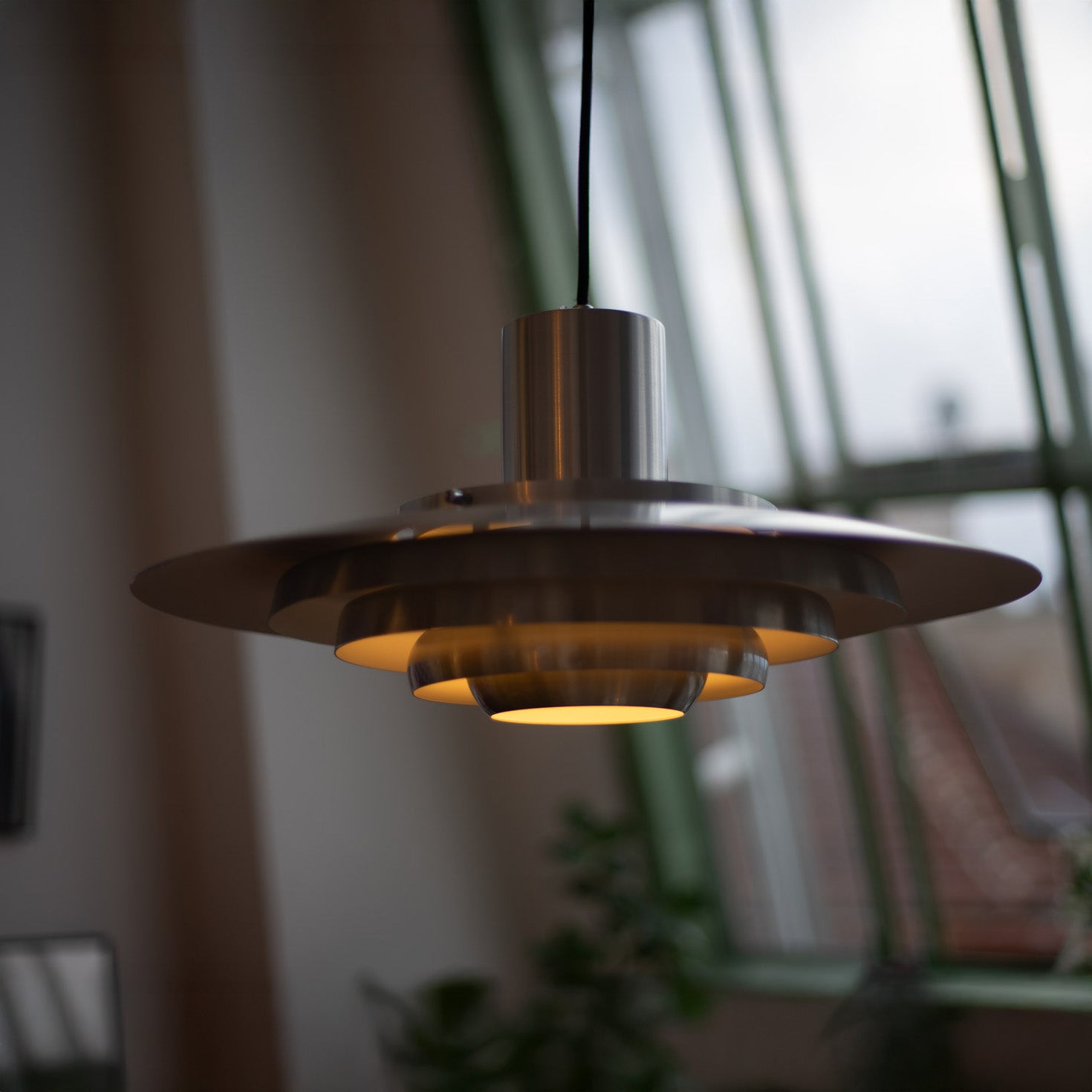 Leuchtende Midcentury-Pendelleuchte aus gebürstetem Metall mit gestufter Form und warmem Licht. Im Hintergrund große grüne Fensterfront mit Blick auf Pflanzen und Dächer einer Stadt.