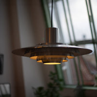 Leuchtende Midcentury-Pendelleuchte aus gebürstetem Metall mit gestufter Form und warmem Licht. Im Hintergrund große grüne Fensterfront mit Blick auf Pflanzen und Dächer einer Stadt.